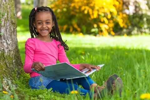 Young girl in a pink shirt reading a book outside and smiling at the camera