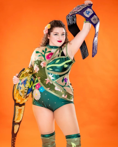the wrestler known as the Unwilting Tatiana wearing a green leotard covered in vines and flowers holding up her two championship belts 