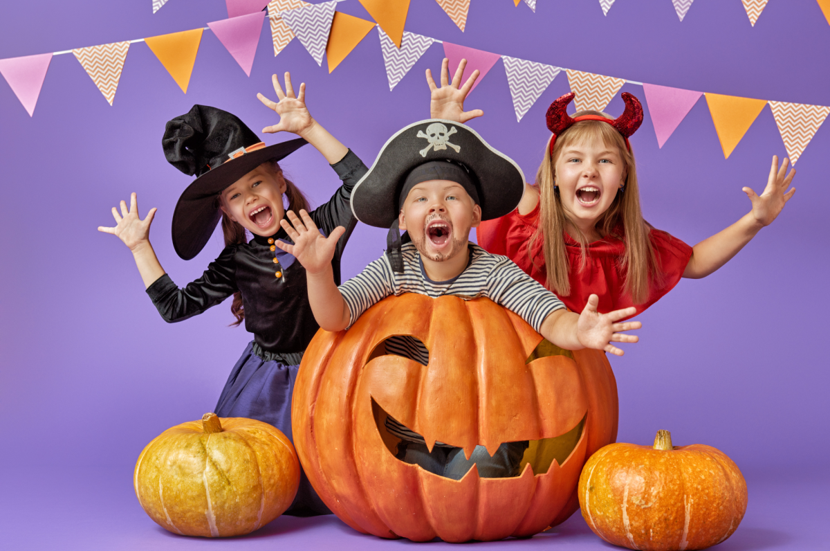 three children in costumes with pumpkins in front a purple background and festive garland